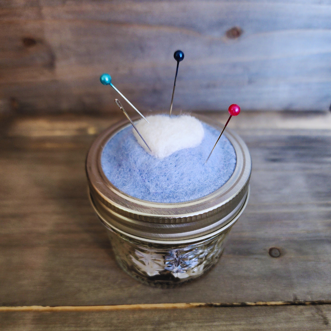 A side-angle view of the completed 4oz jar kit. The jar is filled with assorted white and tan buttons, and the blue wool topper is holding four colorful sewing pins (teal, dark blue, and red).