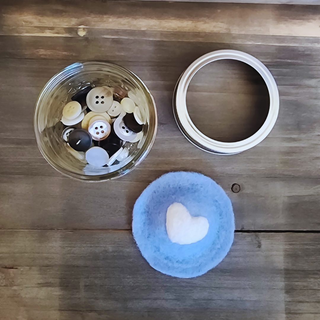 An overhead disassembled view of the blue heart kit, showing the glass jar with buttons, the silver jar ring, and the blue wool heart insert as separate pieces on wood.