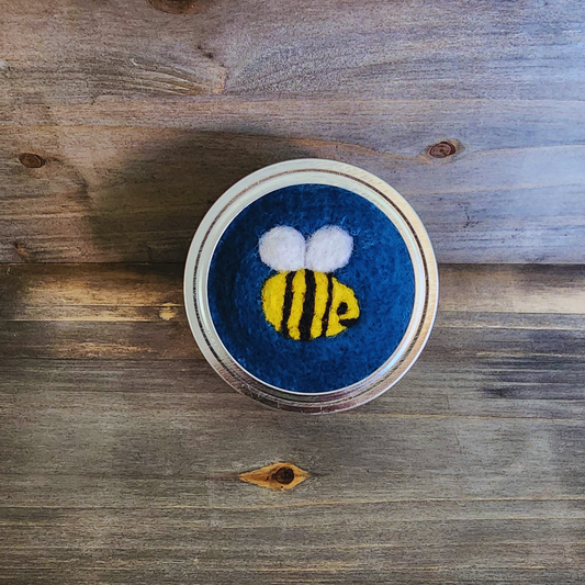 A top-down view of a handcrafted mason jar lid featuring a deep blue needle-felted base. Centered on the blue wool is a small, 3D needle-felted bumblebee with bright yellow and black stripes and two tiny white wings.