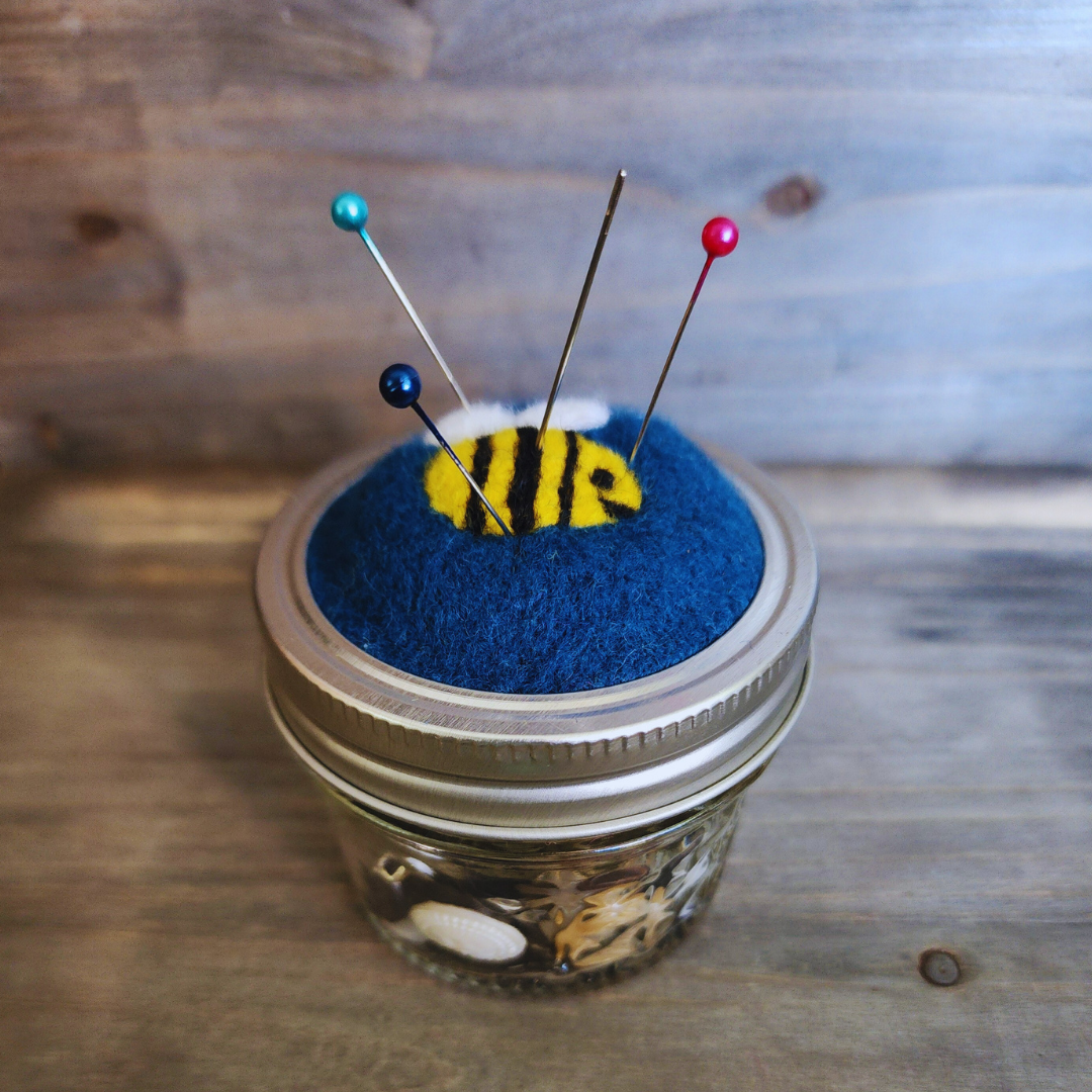 A side-angle view of the bumblebee pincushion on a 4oz jar, with four colorful pins (teal, dark blue, and red) inserted into the blue wool around the felted bee.