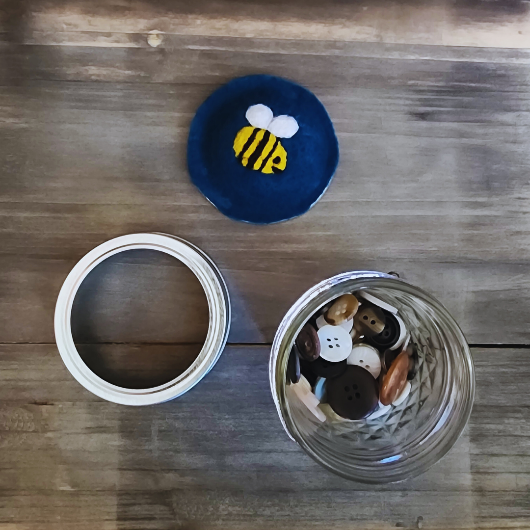 An overhead shot of the disassembled bumblebee kit, showing the blue bee wool insert, the silver jar ring, and the glass jar filled with assorted brown, white, and black buttons.