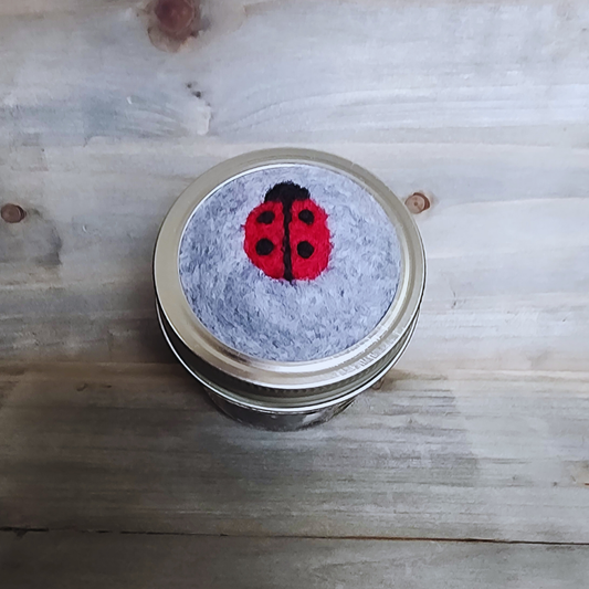 A top-down view of a silver mason jar lid featuring a soft gray needle-felted base. In the center is a bright red needle-felted ladybug with a black head, a black center line, and four black spots.