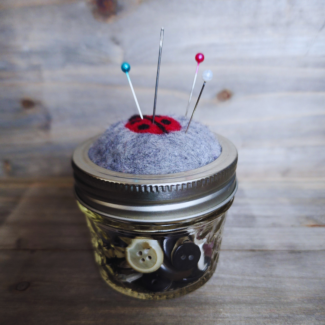 A side-angle view of the ladybug pincushion on a 4oz quilted glass jar filled with buttons. Four sewing pins with colorful heads are inserted into the gray wool base around the ladybug.