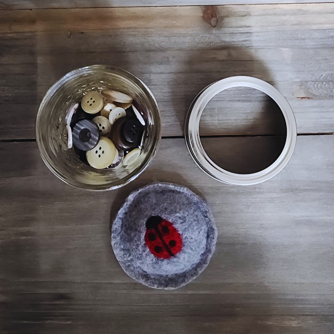 A disassembled view of the ladybug kit on a wooden surface, showing the quilted glass jar filled with buttons, the silver jar ring, and the round gray wool ladybug insert.
