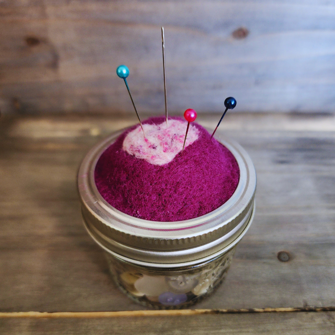 A side-angle view of the magenta heart pincushion holding a needle and three pins with teal, pink, and dark blue heads.