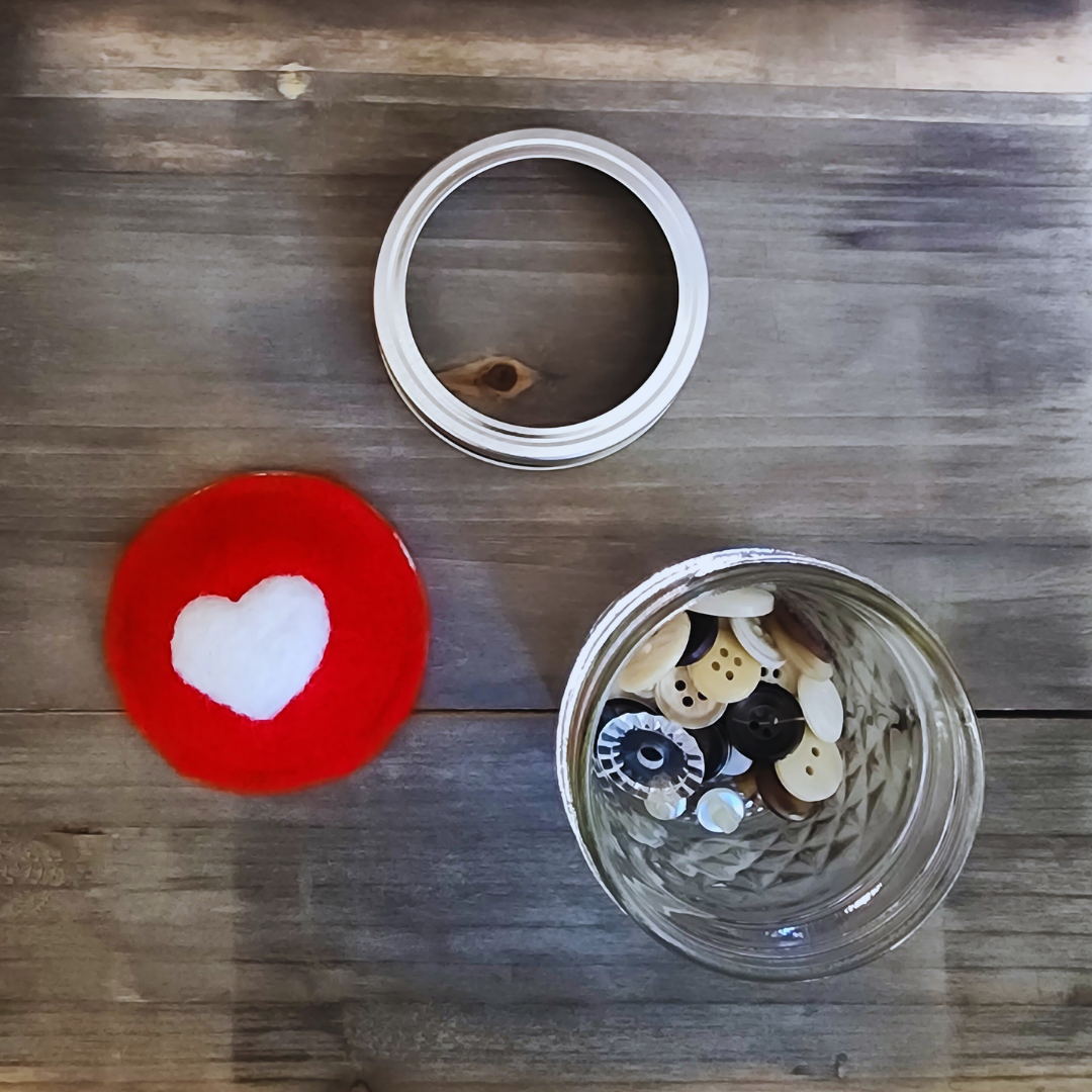 A disassembled kit view showing the red heart wool insert, silver ring, and glass jar with buttons on a wooden background.