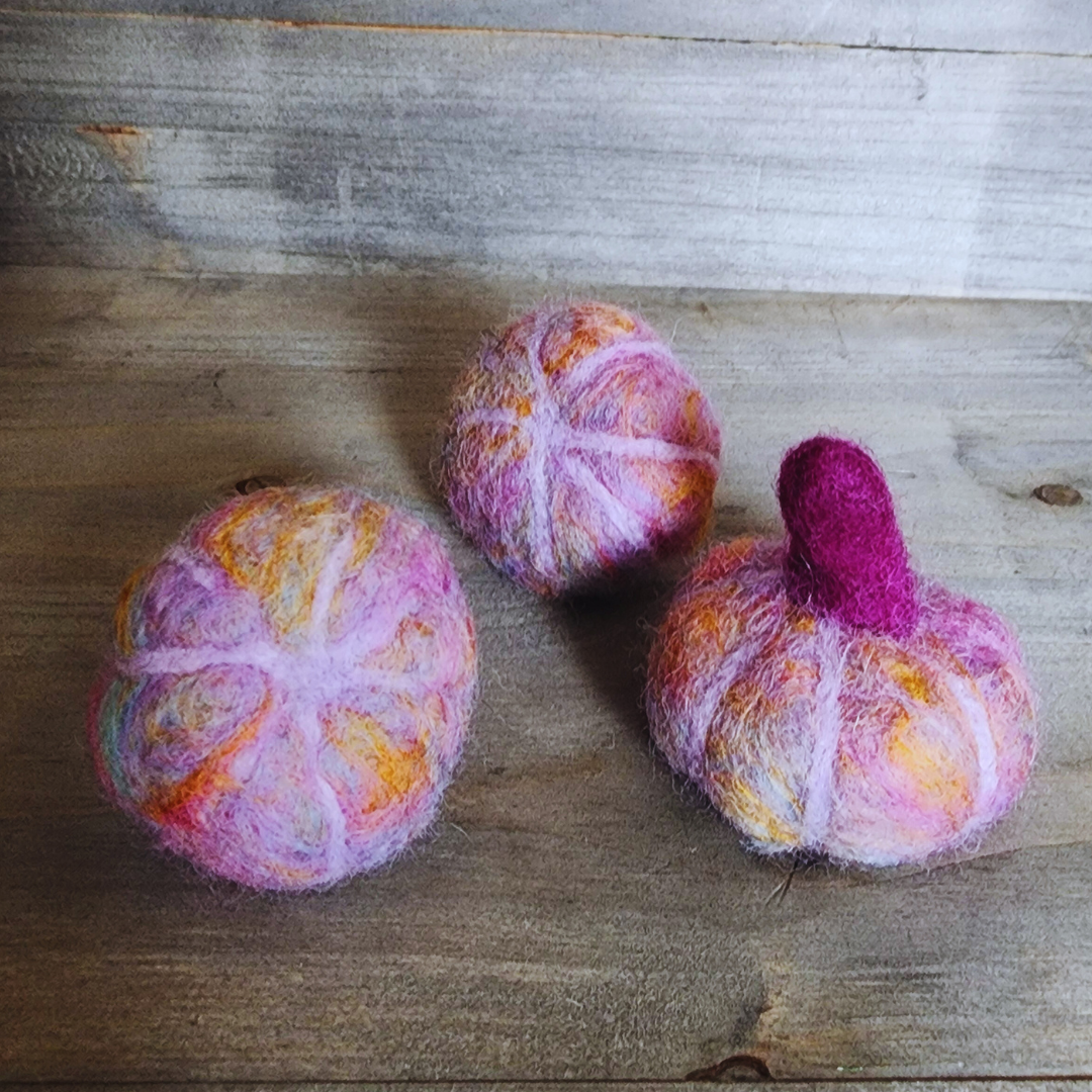 Three handmade felted pumpkins in shades of purple and pink with orange accents and one with a purple felted stem, arranged in a triangular pattern on a wooden surface.