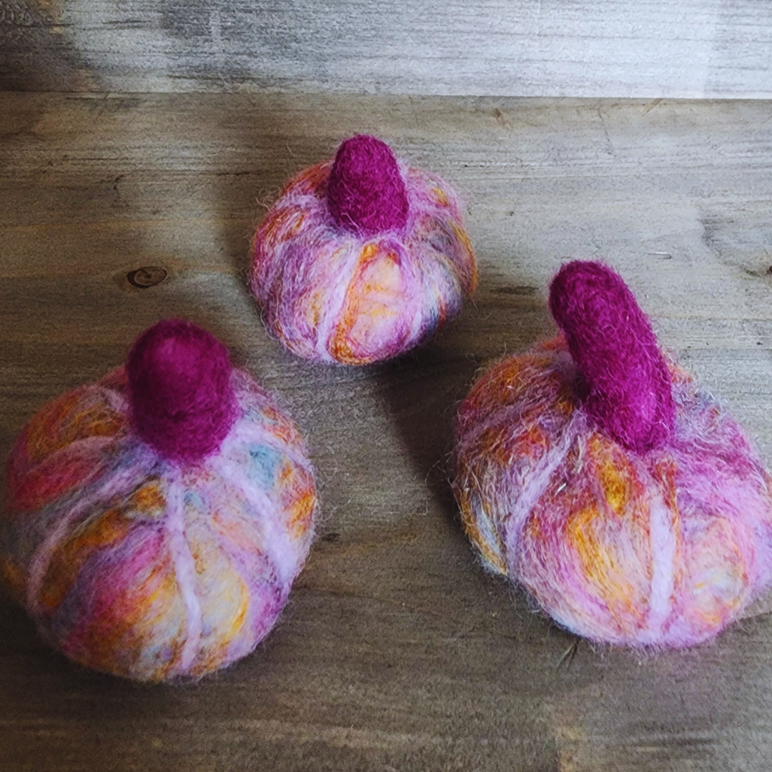 Three handmade felted pumpkins in shades of purple, pink, and orange, each with a vibrant magenta felted stem, arranged in a triangular pattern on a wooden surface.
