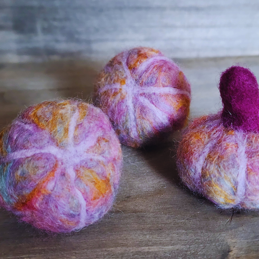 Three handmade felted pumpkins in shades of purple, pink, and orange, one with a prominent magenta felted stem, arranged in a triangular pattern on a wooden surface.