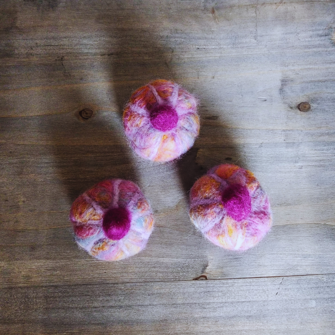 An overhead view of three handmade felted pumpkins in shades of purple, pink, and orange, each with a prominent magenta felted stem, arranged in a triangular pattern on a wooden surface.