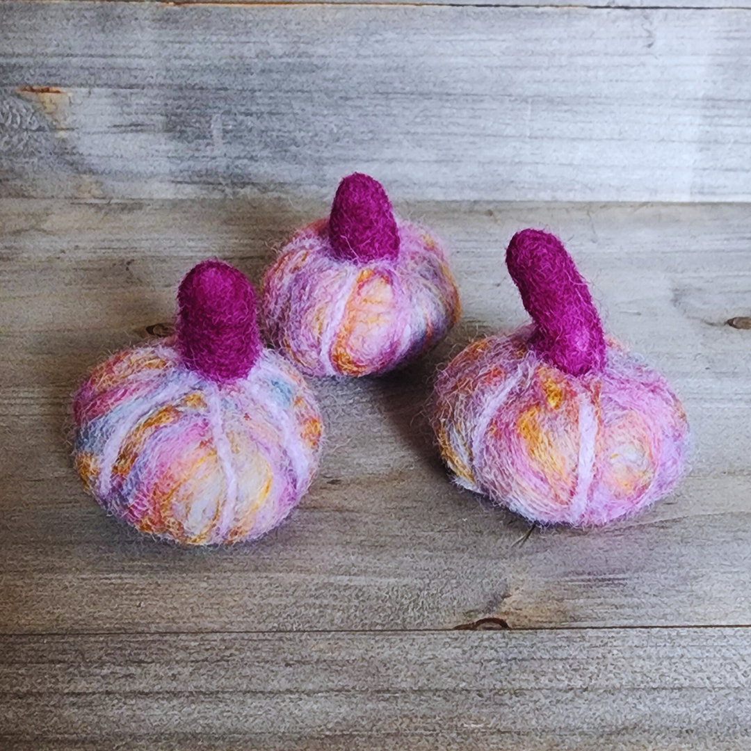 Three handmade felted pumpkins in shades of purple, pink, and orange, each with a magenta felted stem, arranged in a triangular pattern on a wooden surface.