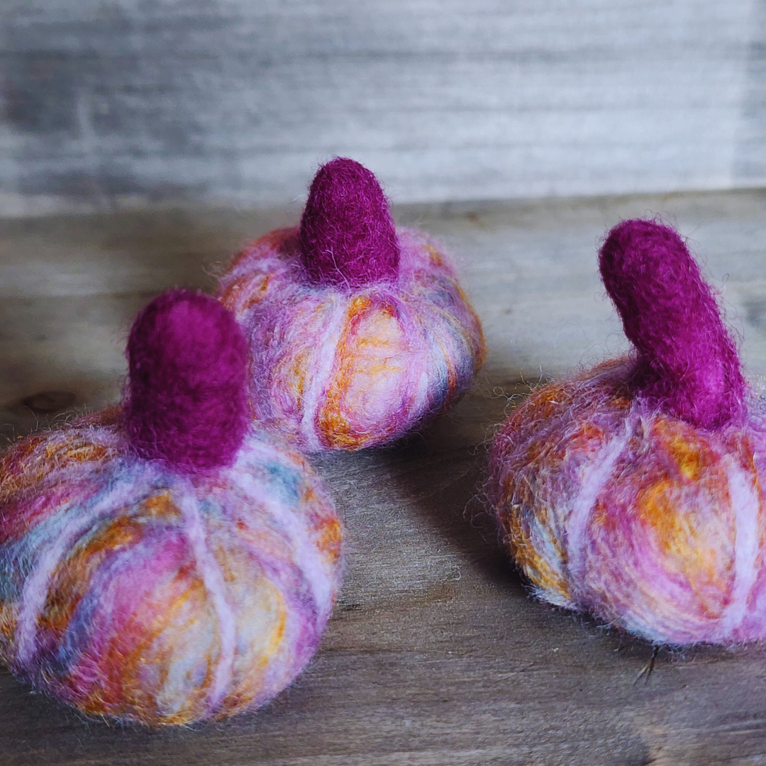 A close-up of three handmade felted pumpkins in shades of purple, pink, and orange, each with a magenta felted stem, showing their textures and vibrant colors on a wooden background.