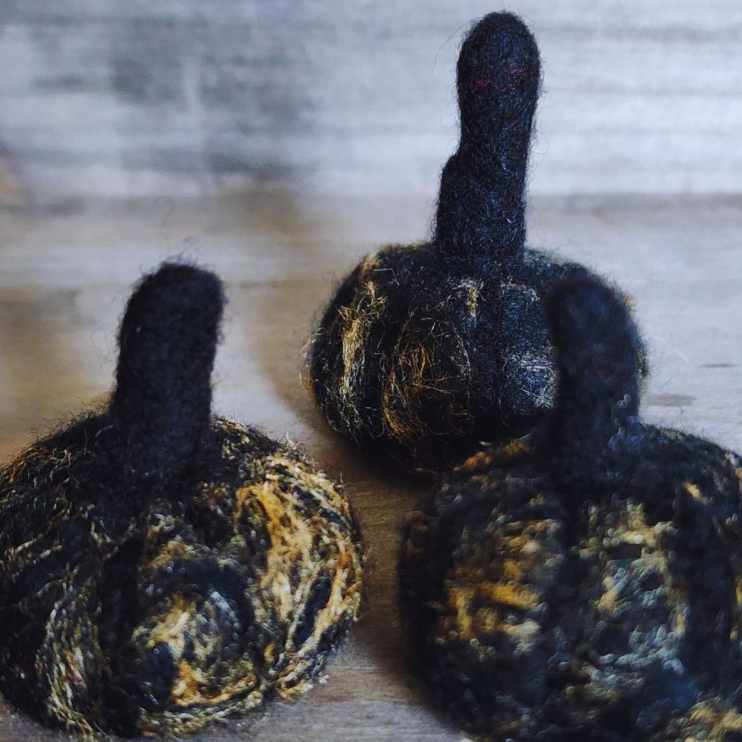 A close-up of three handmade felted black pumpkins with subtle gold flecks and dark stems, arranged on a wooden surface.