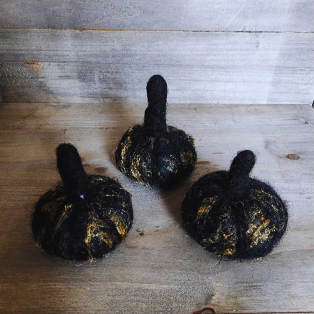  Three handmade felted black pumpkins with subtle gold flecks and dark stems, arranged in a triangular pattern on a wooden surface.
