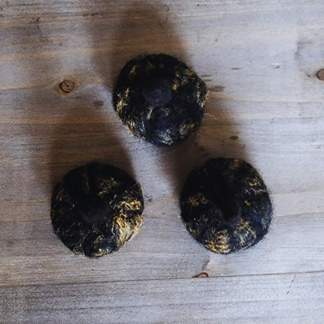 An overhead view of three handmade felted black pumpkins with subtle gold flecks, showing their unique textures, arranged on a wooden surface.