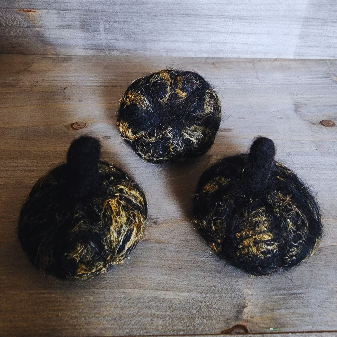 Three handmade felted black pumpkins with prominent gold flecks and dark stems, arranged in a triangular pattern on a wooden surface.