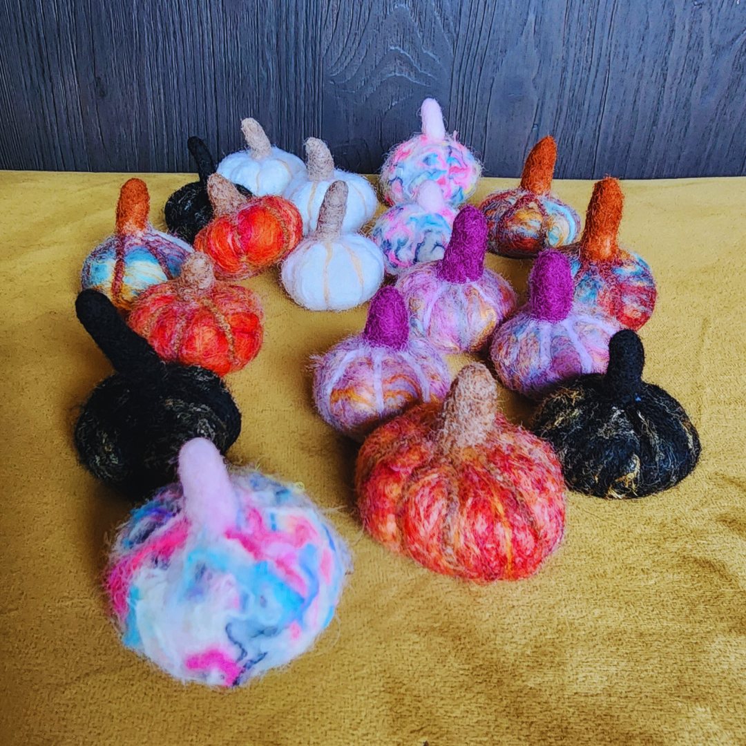 A large group of assorted handmade felted pumpkins in various colors (black, white, red, orange, pink, purple, multicolor), arranged on a yellow fabric surface against a wooden backdrop.