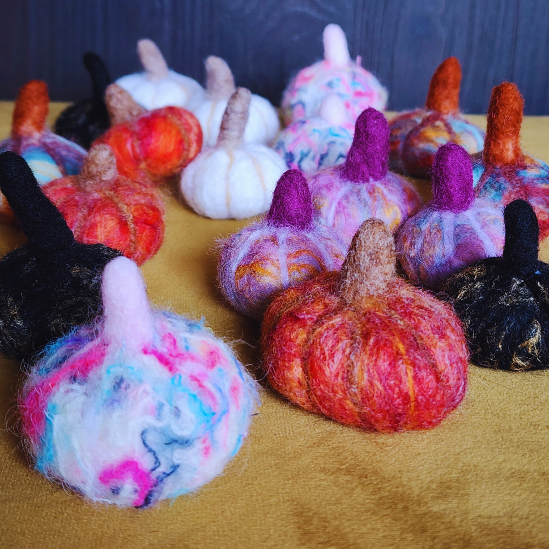 A cluster of assorted handmade felted pumpkins in various colors (black, white, red, orange, pink, purple, multicolor), arranged on a yellow fabric surface with a wooden backdrop.