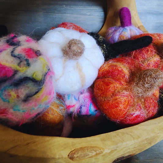 A wooden bowl filled with various handmade felted pumpkins in assorted colors, including white, black, orange, and neon multicolor, showing their unique stems and textures.