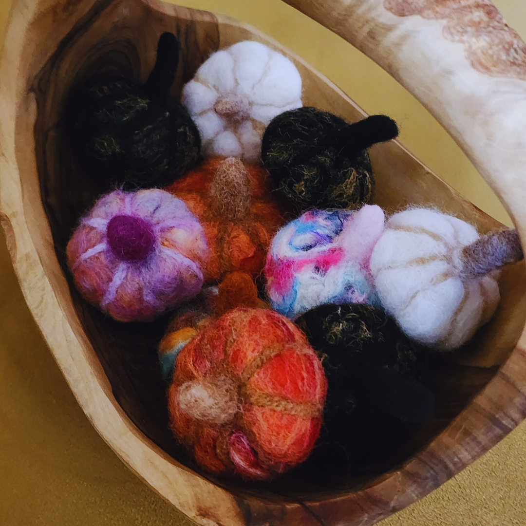 An overhead view of a wooden bowl filled with various handmade felted pumpkins in assorted colors, including white, black, orange, pink, purple, and neon multicolor, showing their unique stems and textures.
