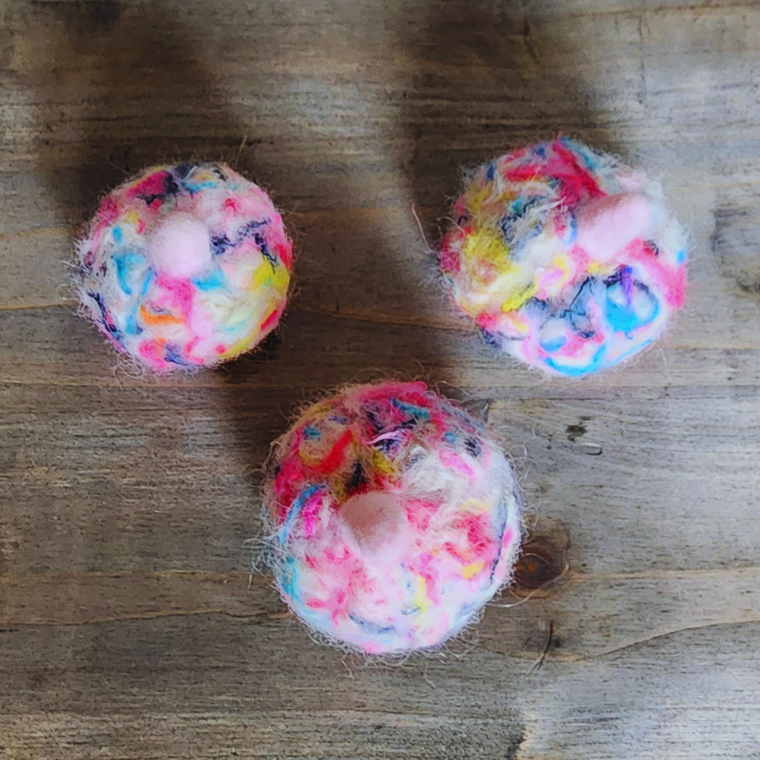 Three handmade felted pumpkins in bright neon multicolor (pink, yellow, blue, purple) with pink stems, viewed from above on a wooden surface.
