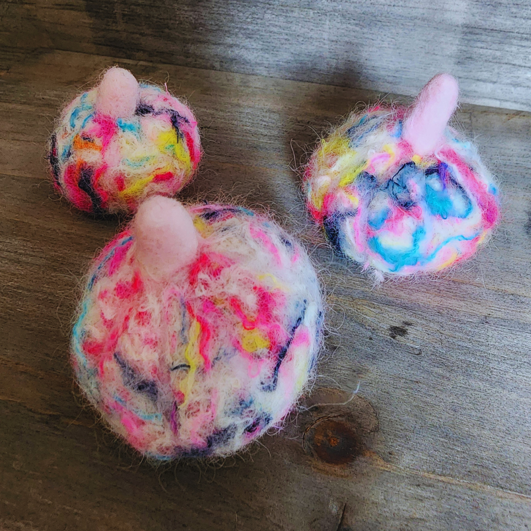 Three handmade felted pumpkins in bright neon multicolor (pink, yellow, blue, purple) with pink stems, arranged in a triangular pattern on a wooden surface.