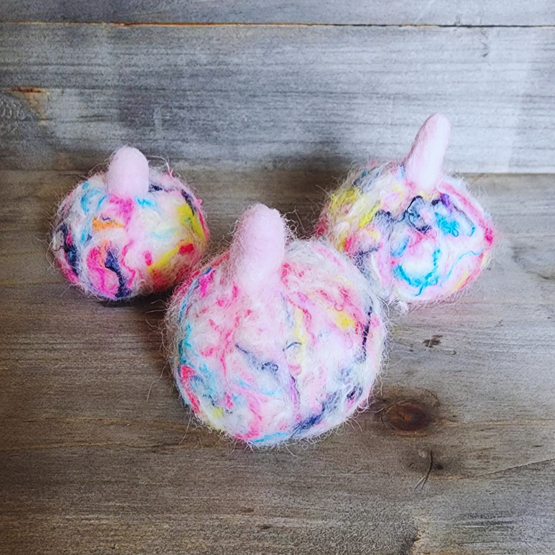 Three handmade felted pumpkins in bright neon multicolor (pink, yellow, blue, purple) with pink stems, arranged in a triangle on a wooden surface.