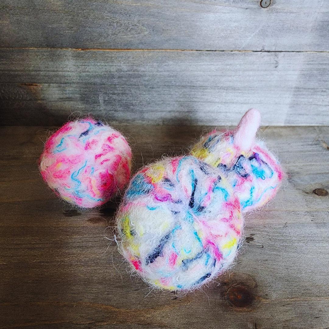 Three handmade felted pumpkins in bright neon multicolor (pink, yellow, blue, purple) with pink stems, arranged in a triangular pattern on a wooden surface.