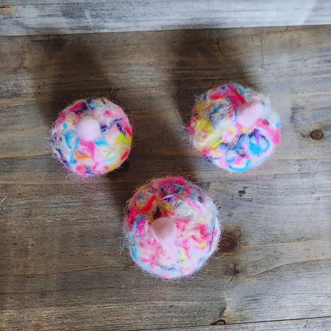 Three handmade felted pumpkins in bright neon multicolor (pink, yellow, blue, purple) with pink stems, arranged in a triangle on a wooden surface.