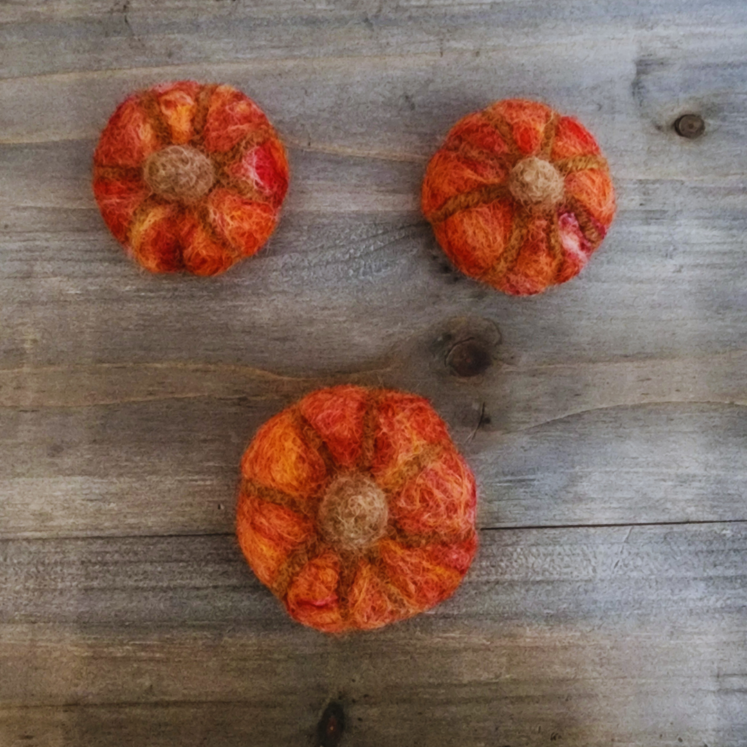Three handmade felted pumpkins in shades of orange and red with brown felted stems, arranged in a triangular pattern on a wooden surface.