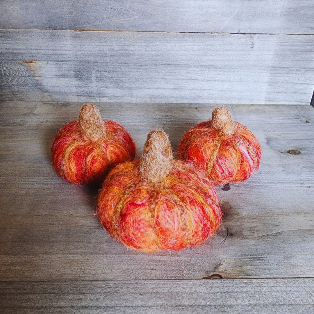 Three handmade felted pumpkins in shades of orange and red with brown felted stems, arranged in a triangular pattern on a wooden surface.