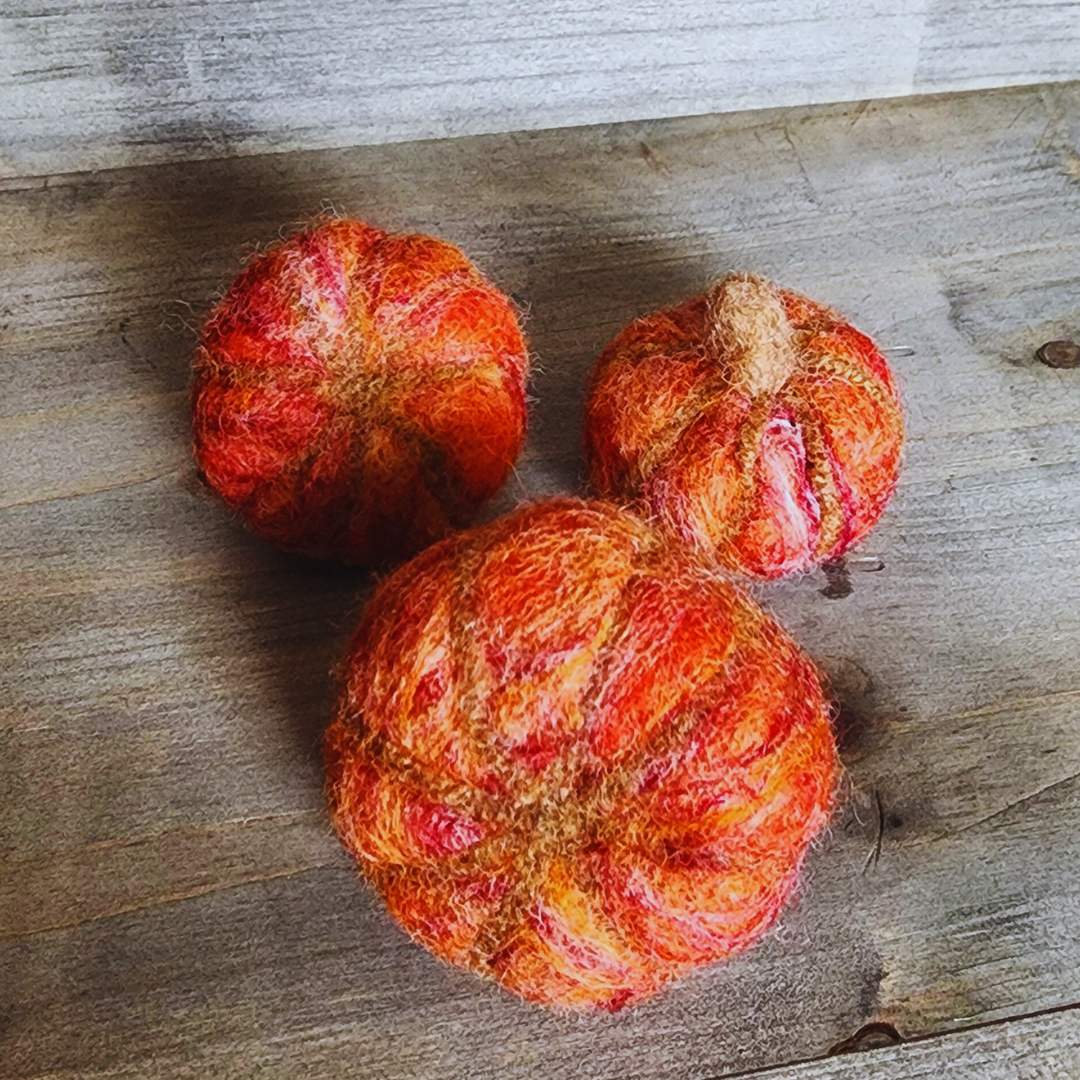 Three handmade felted pumpkins in shades of orange and red with brown felted stems, arranged in a triangular pattern on a wooden surface.