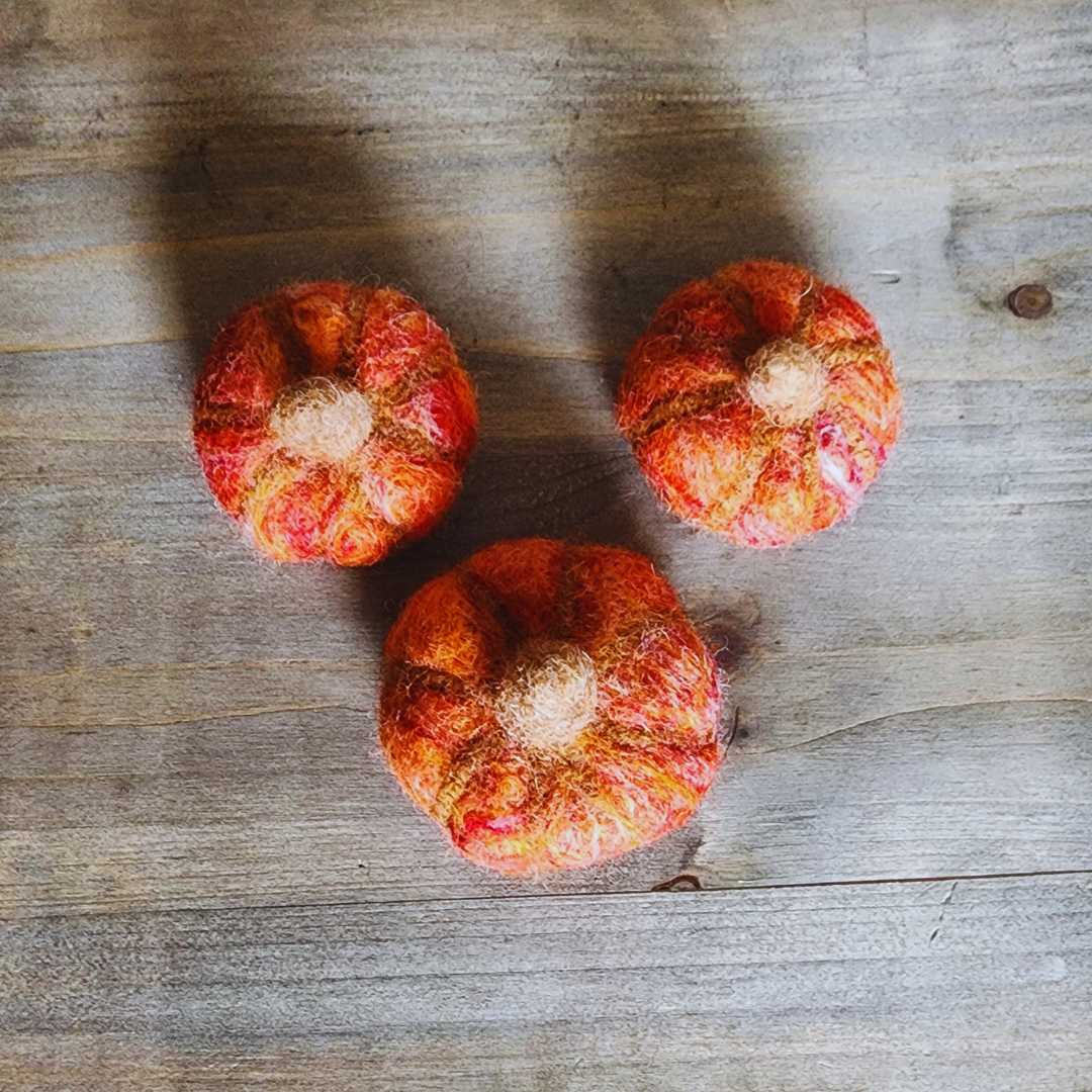 Three handmade felted pumpkins in shades of orange and red with brown felted stems, arranged in a triangular pattern on a wooden surface.