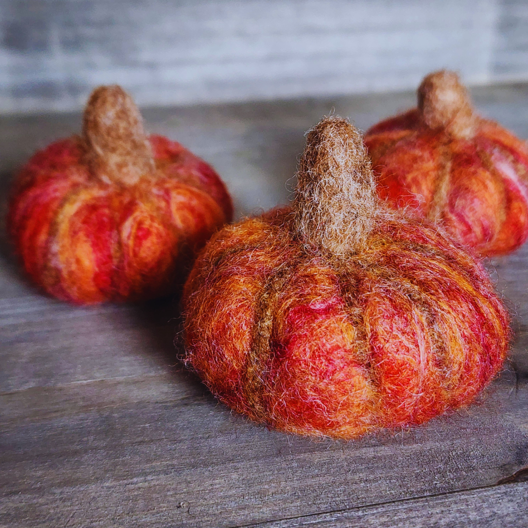 A close-up of three handmade felted pumpkins in shades of orange and red with brown felted stems, showing their textured surface on a wooden background.