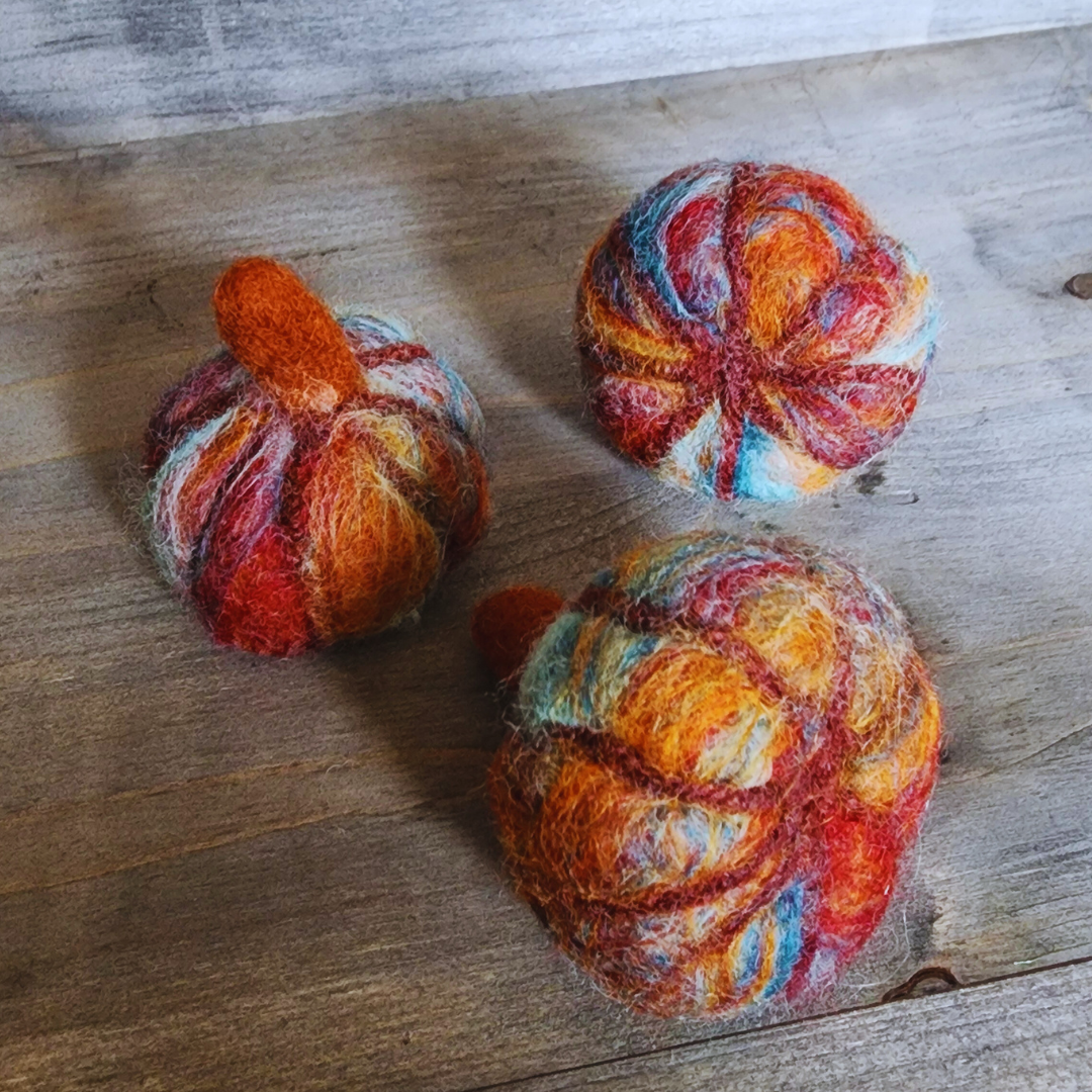 Three handmade felted pumpkins in warm Sedona-inspired colors (orange, red, blue, and brown) with orange felted stems, arranged in a triangular pattern on a wooden surface.