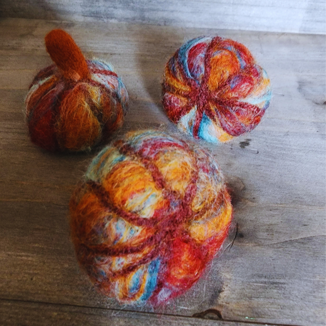 Three handmade felted pumpkins in warm Sedona-inspired colors (orange, red, blue, and brown) with orange felted stems, arranged in a triangular pattern on a wooden surface.