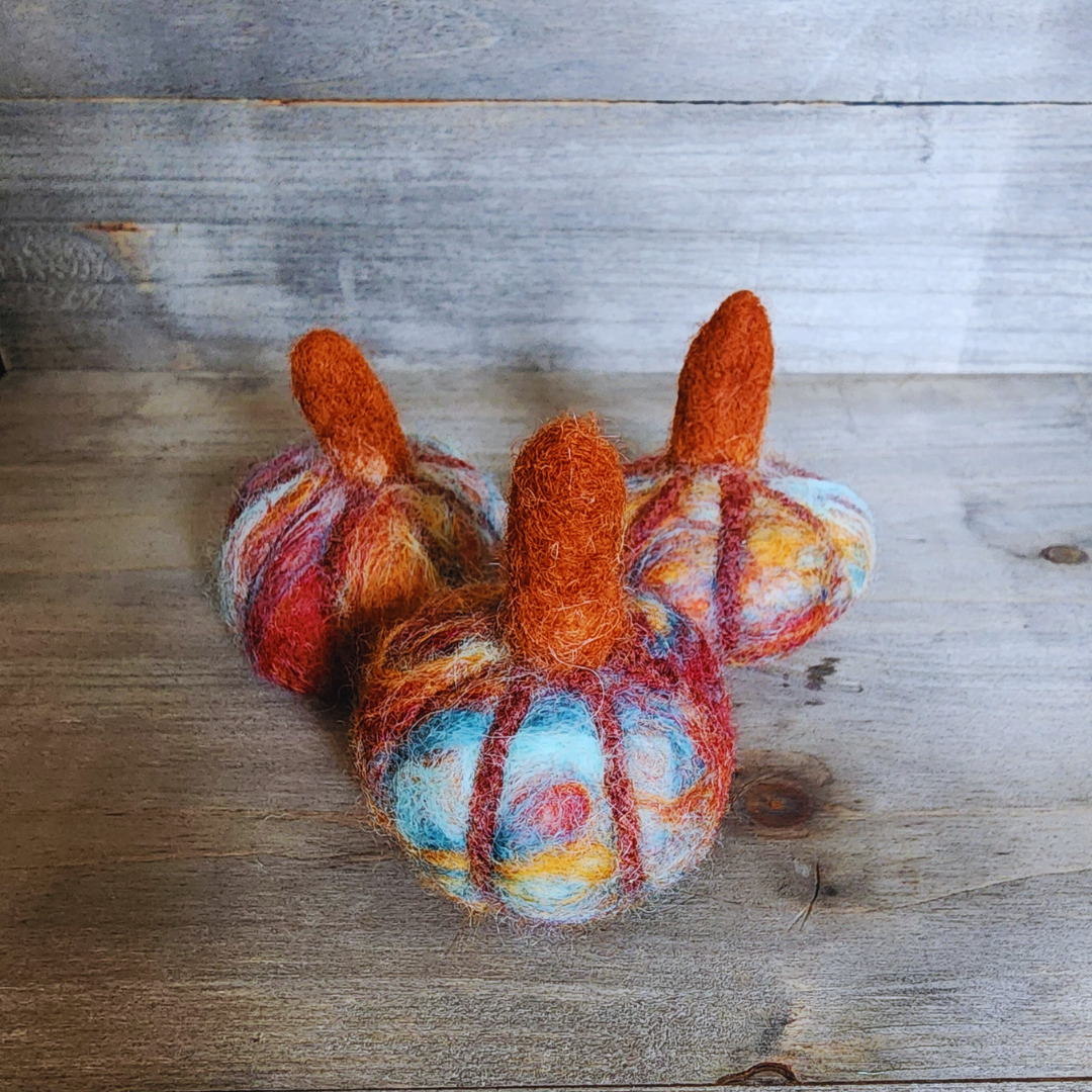 Three handmade felted pumpkins in warm Sedona-inspired colors (orange, red, blue, and brown) with prominent orange felted stems, arranged in a triangular pattern on a wooden surface.