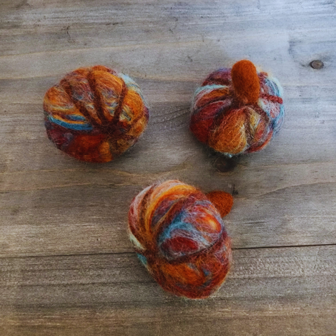 Three handmade felted pumpkins in warm Sedona-inspired colors (orange, red, blue, and brown) with orange felted stems, arranged in a triangular pattern on a wooden surface.