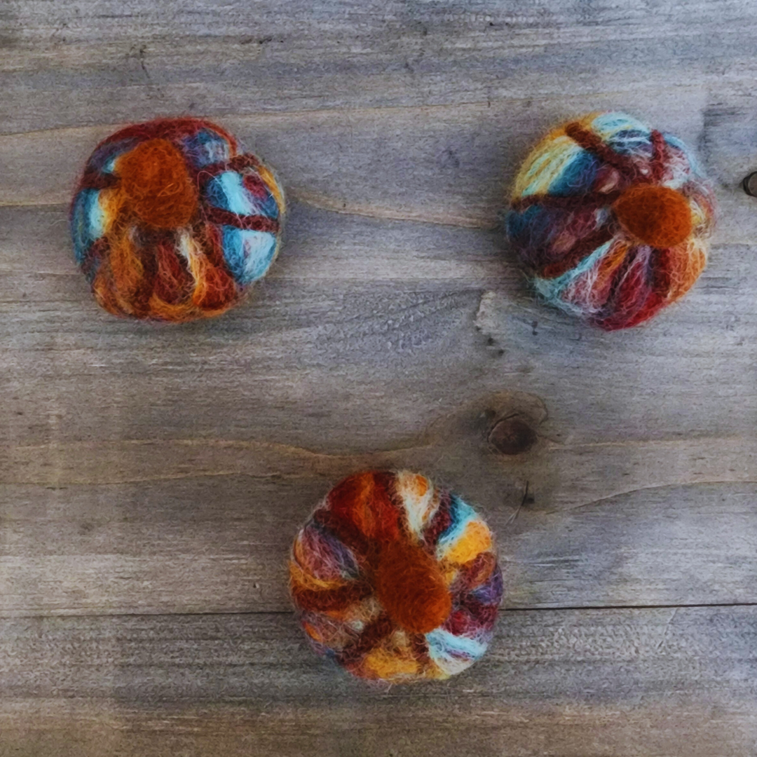 An overhead view of three handmade felted pumpkins in warm Sedona-inspired colors (orange, red, blue, and brown) with orange felted stems, arranged in a triangular pattern on a wooden surface.