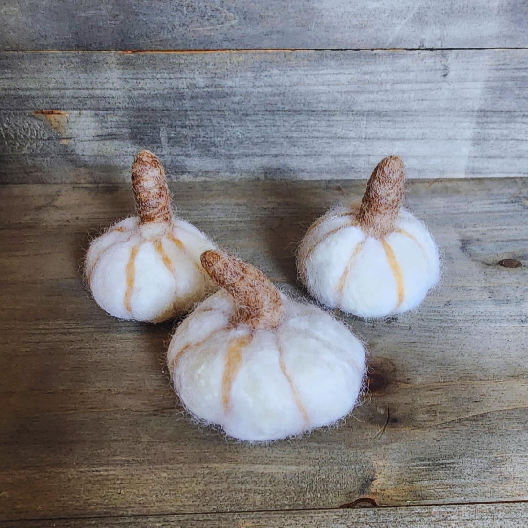 Three handmade felted white pumpkins, each with a brown felted stem and subtle vertical orange stripes, arranged in a triangular pattern on a wooden surface.