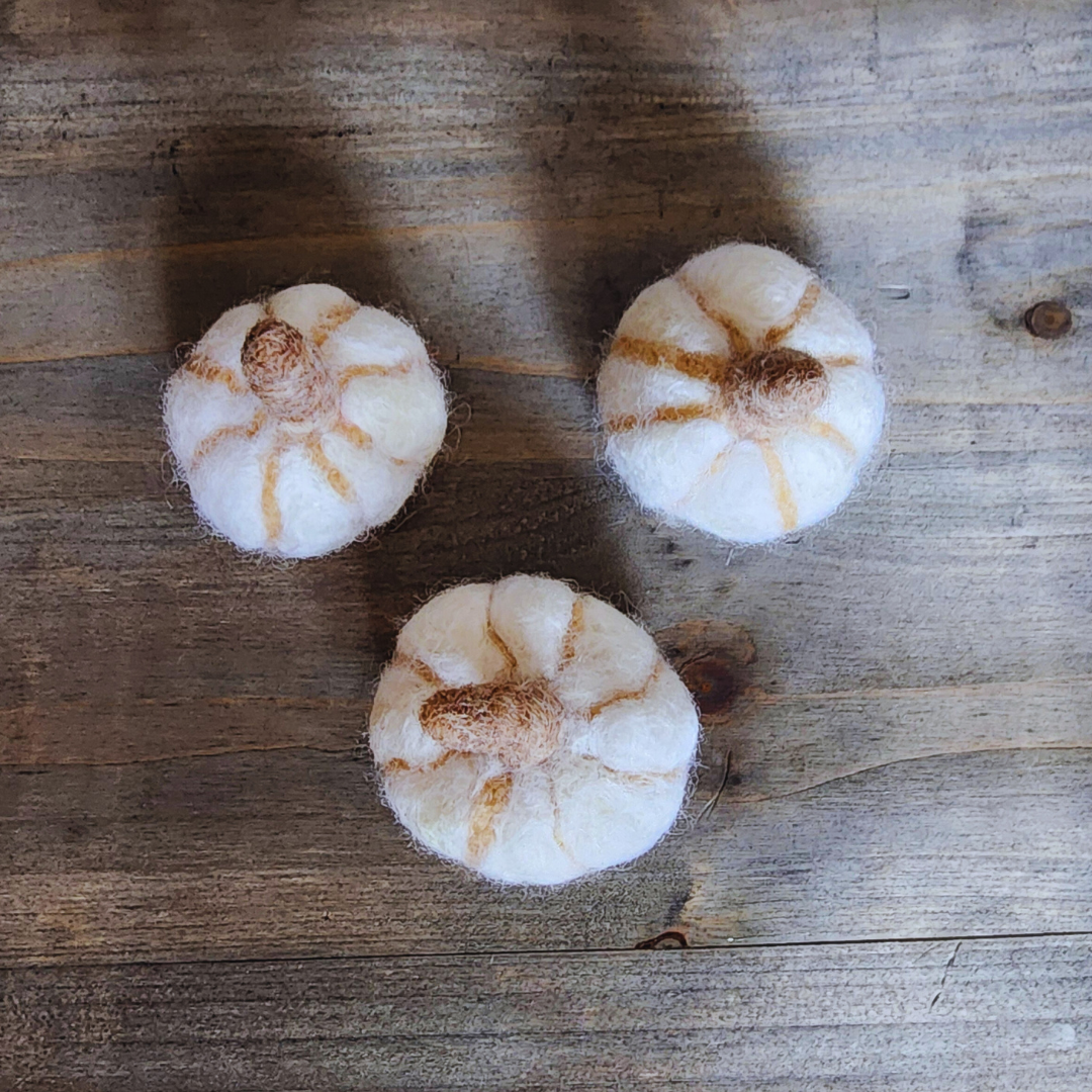 An overhead view of three handmade felted white pumpkins, each with a brown felted stem and subtle vertical orange stripes, arranged in a triangular pattern on a wooden surface.