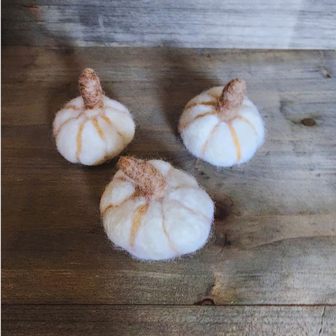 Three handmade felted white pumpkins, each with a brown felted stem and subtle vertical orange stripes, arranged in a triangular pattern on a wooden surface.