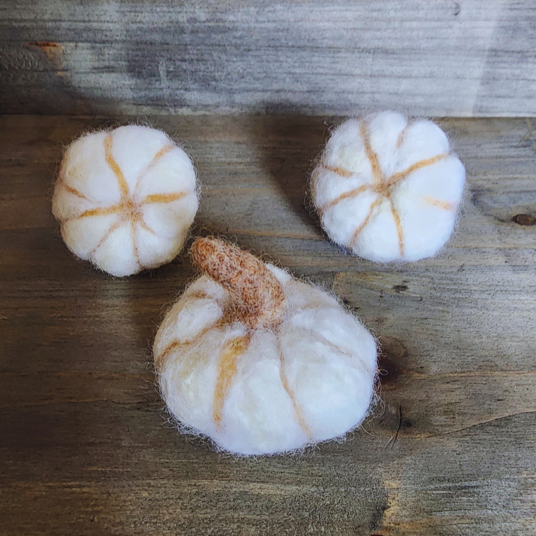 Three handmade felted white pumpkins, each with a prominent brown felted stem and subtle vertical orange stripes, arranged in a triangular pattern on a wooden surface.