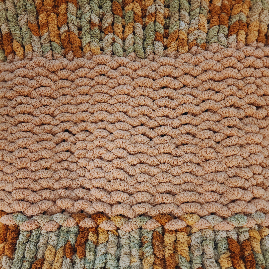 A close-up overhead view of a handmade chunky knit blanket with a single stitch pattern, showing a solid peach-colored horizontal band separating two sections of multicolor yarn in desert sand tones (orange, brown, light grey).