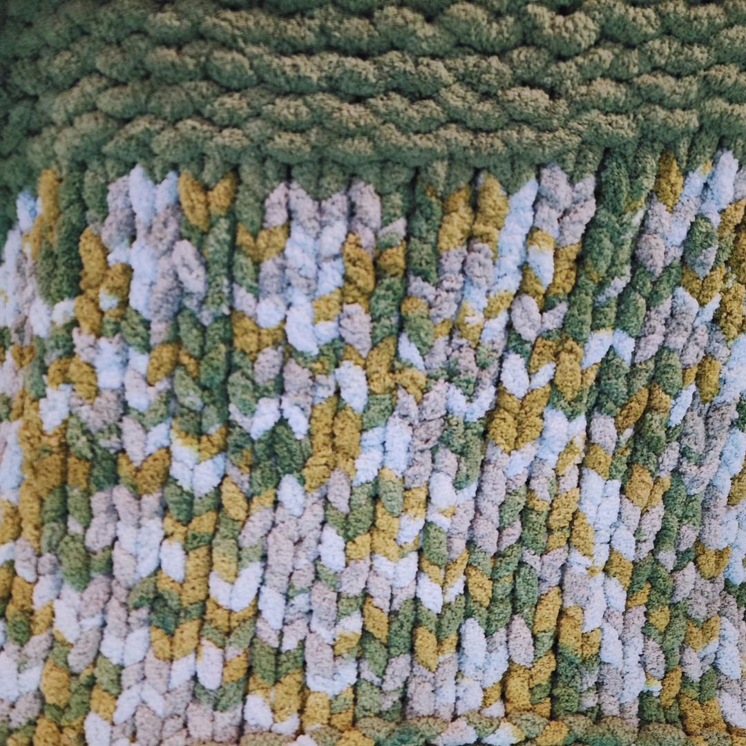 A close-up overhead view of a handmade chunky knit blanket with a single stitch pattern, showing a solid green row at the top transitioning to a textured multicolor pattern of green, yellow, and white.