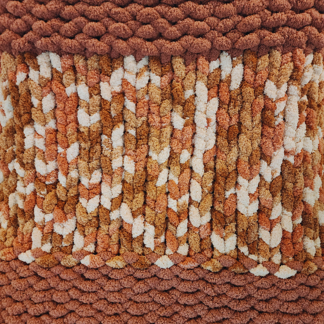 A close-up overhead view of a handmade chunky knit blanket with a single stitch pattern, showing a solid dark reddish-brown border at the top and bottom, framing a central section with a textured multicolor pattern of orange, brown, and white.