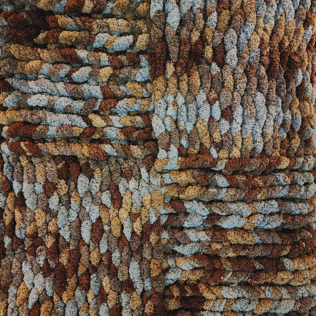 A close-up overhead view of a handmade chunky knit blanket with a checkerboard pattern, showing squares of two different multicolor blends of brown, gray, and white yarn.