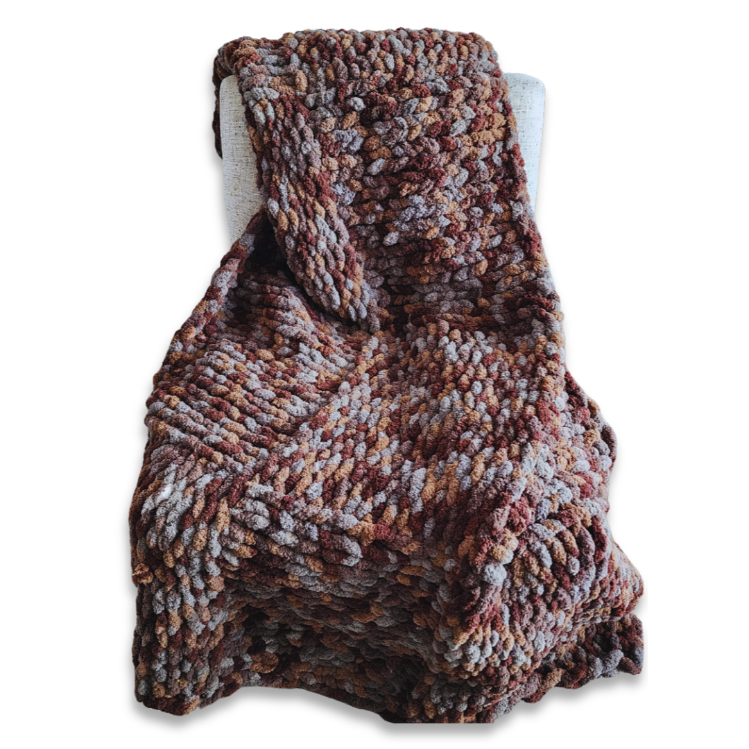 A handmade chunky knit blanket in a brown and gray multicolor yarn blend, draped over the back of a light-colored upholstered chair against a white background.