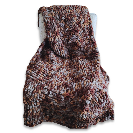 A handmade chunky knit blanket in a brown and gray multicolor yarn blend, draped over the back of a light-colored upholstered chair against a white background.