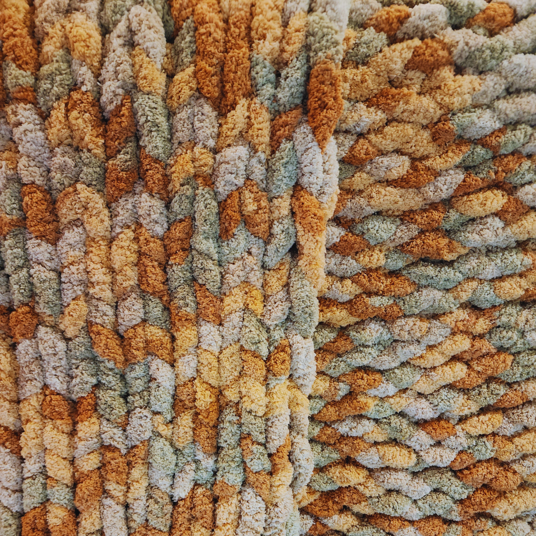 A close-up overhead view of a handmade chunky knit blanket, showing its soft, textured surface and multicolor pattern in earthy tones of orange, brown, and light green.